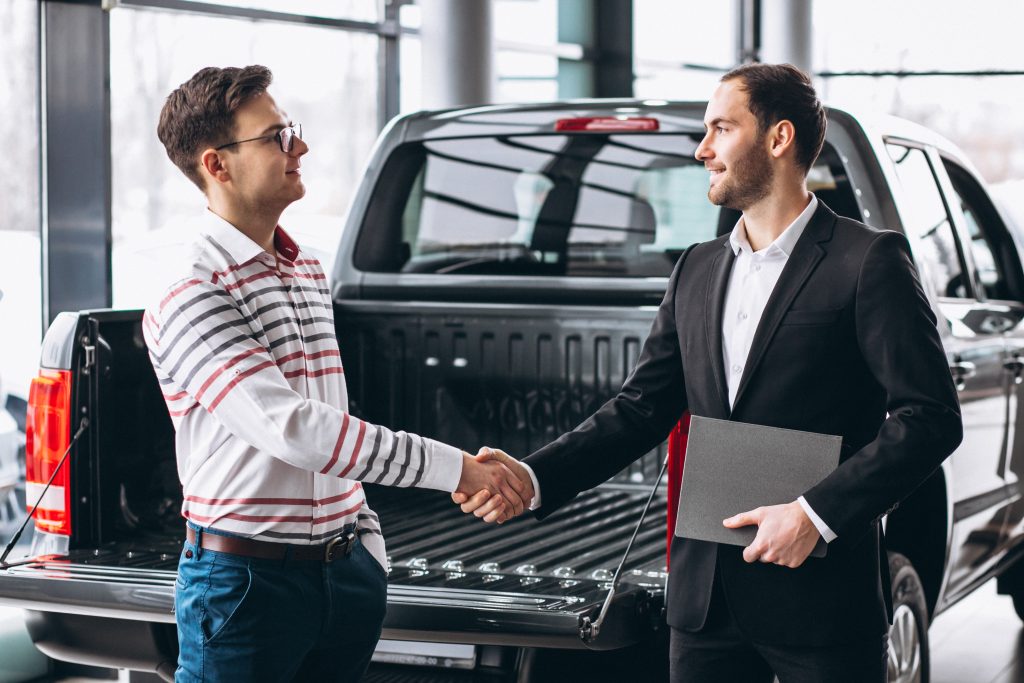 Two men shaking hands in a car dealership next to the open bed of a black pickup truck.