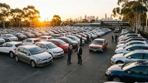 Two men shake hands in front of a large car dealership lot with the Melbourne skyline in the background at sunset.